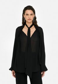 Blouse noire en tissu léger, avec un décolleté en trou de serrure, un détail à nouer et des manches longues ornées de volants discrets aux épaules.