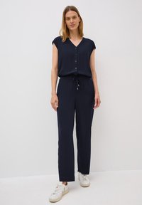 Marineblauw jumpsuit met een V-hals, korte mouwen, knopen aan de voorkant en een elastische taille. Heeft rechte benen en een strikdetail. Gekocht met witte sneakers.