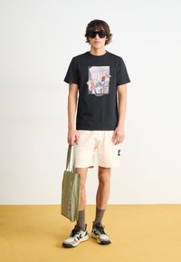 Svart t-skjorte med et fargerikt grafisk trykk, kombinert med lyse shorts og en grønn tote bag. Modellen har på seg oversized solbriller og sneakers.