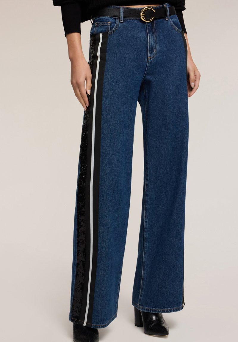 Szerokie dżinsowe spodnie w ciemnoniebieskim kolorze z bocznymi paskami w czerni i srebrze. Posiadają teksturowany czarny panel oraz dopasowany czarny pasek z złotą klamrą.