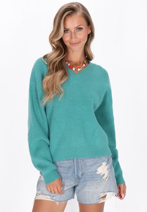 Pull en tricot turquoise avec un col en V, des poignets côtelés et des détails sur les coutures latérales, associé à un short en denim déchiré.