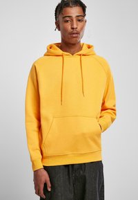 Sudadera amarilla con bolsillo tipo canguro, capucha con cordón y puños acanalados. Tejido suave y corte holgado. Se lleva con pantalones oscuros.