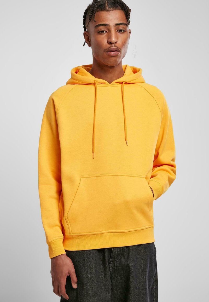 Sudadera amarilla con bolsillo tipo canguro, capucha con cordón y puños acanalados. Tejido suave y corte holgado. Se lleva con pantalones oscuros.