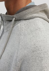 Sudadera con capucha gris de punto con cierre de cremallera, cuello texturizado en contraste y cordones. Presenta una tela suave y jaspeada y hombros sin costuras.