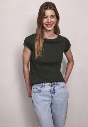 Junge Frau mit langen hellbraunen Haaren, trägt ein dunkelgrünes kurzärmeliges Shirt und hellblaue Jeans, lächelt mit den Händen hinter dem Rücken.