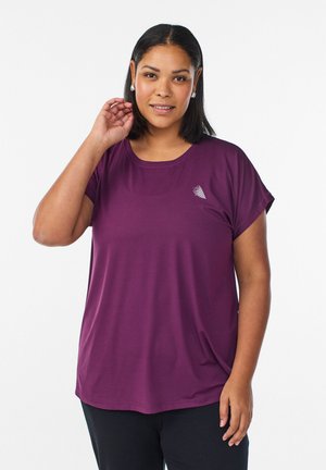 Woman with shoulder-length dark hair wearing silver hoop earrings, a loose purple t-shirt with logo, and black pants, standing against white background.