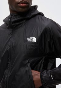 Άνδρας που φοράει μαύρο μπουφάν North Face Windwall με φερμουάρ μπροστά και χέρι στην τσέπη, εμφανίζεται από το στήθος μέχρι το λαιμό.