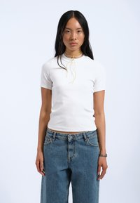 Hvid kortærmet skjorte med en tætsiddende design, parret med lyseblå brede denimjeans. Accenteret med lagdelte halskæder, der har et gult slips.