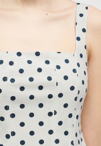 Γυναίκα που φοράει λευκό αμάνικο φόρεμα με μαύρες πουά, που δείχνει την περιοχή του ντεκολτέ και τους ώμους.