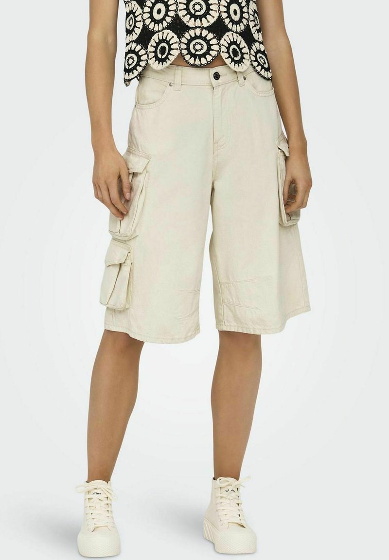 Shorts cargo beige avec plusieurs poches latérales, fabriqués dans un tissu léger. Associé à un haut à motifs noir et blanc et des baskets blanches.