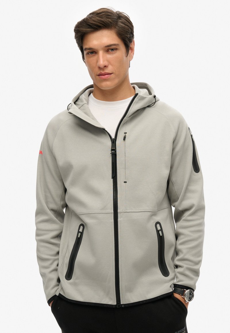 Chaqueta con capucha de color gris claro, fabricada en tejido suave, con una cremallera frontal, bolsillos con cremallera negros y capucha con cordón ajustable.