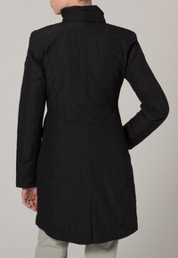 Veste noire imperméable avec un col haut, coupe ajustée et coutures contrastées. Elle dispose de poches latérales et d'un logo sur le dos.