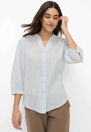 Junge Frau, die ihre Haare richtet, trägt eine hellblaue, gestreifte Bluse mit Knöpfen und 3/4-Ärmeln sowie eine braune Hose vor weißem Hintergrund.