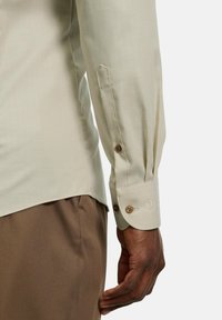 Die Hand eines Mannes liegt entspannt zur Seite, während er ein beiges, geknöpftes Hemd und braune Hose vor einem weißen Hintergrund trägt.