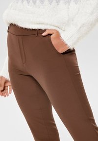 Bruna figursydda byxor med en slät yta, smal passform och bältesöglor, kombinerat med en vit fluffig tröja på bilden.