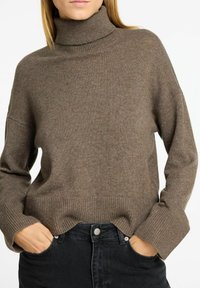Brauner Strickrollkragenpullover mit lockerem Schnitt, gerippten Bündchen und Saum, kombiniert mit schwarzen Jeans. Weiche Textur, keine sichtbaren Muster.