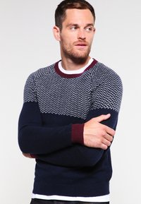 Navy blue sweater with a white zigzag pattern on the chest. Ribbed maroon cuffs and collar. Soft knit fabric, fitted design.