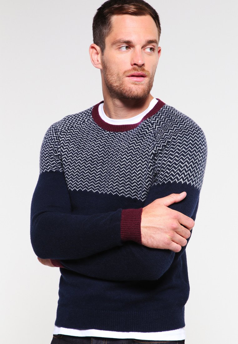 Navy blue sweater with a white zigzag pattern on the chest. Ribbed maroon cuffs and collar. Soft knit fabric, fitted design.