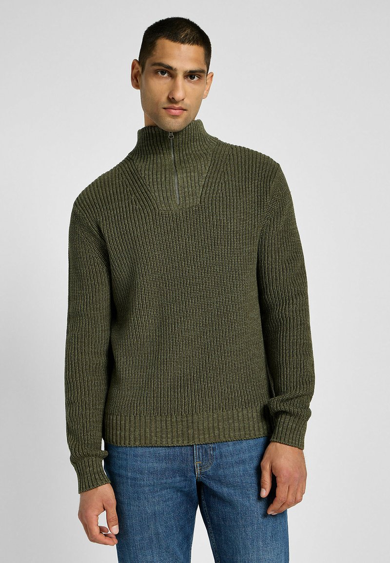 Tummanvihreä ribbiknitti neulepaita korkealla kauluksella ja puolivetoketjulla, jossa on pitkät hihat ja teksturoitu kuvio. Yhdistetty sinisiin farkkuihin.