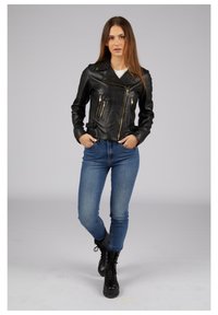 Schwarze Leder-Bikerjacke mit goldenen Reißverschlüssen, ausgestattet mit einem strukturierten Strickkragen, kombiniert mit hellblauen Jeans und schwarzen Schnürstiefeln.