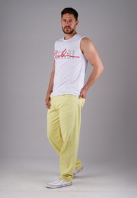 White sleeveless shirt with red and grey text, paired with loose yellow pants and white sneakers. Simple design and casual fit.