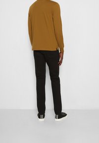 Langärmliges Senfshirt mit Rundhalsausschnitt, kombiniert mit slim-fit schwarzen Hosen und schwarzen Sneakers mit weißen Streifen an der Seite.