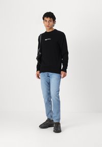 Juodadrabio megztinis su apvaliu kaklu, su baltu logotipu ant krūtinės, derinamas su šviesiai mėlynomis džinsomis ir juodais sportbačiais su tekstūruotu detalizavimu.