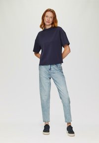 Navy-blaues Kurzarm-Oberteil mit hohem Kragen, kombiniert mit hellblauen, taillierten Jeans und dunklen Sneakers, vor einem neutralen Hintergrund.