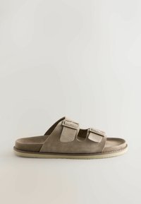Sandale mule en daim beige avec deux boucles réglables, design à bout ouvert et semelle texturée. Présente une esthétique lisse et minimaliste.