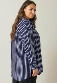 Camisa de rayas de manga larga en azul marino y blanco, con puños abotonados, dobladillo curvado y un corte relajado con detalle en el yugo trasero.