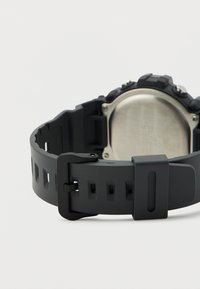 Μαύρο ψηφιακό ρολόι χειρός με ρυθμιζόμενο λουράκι από καουτσούκ και μεταλλική πίσω πλάκα, εμφανιζόμενο από πίσω σε λευκό φόντο.