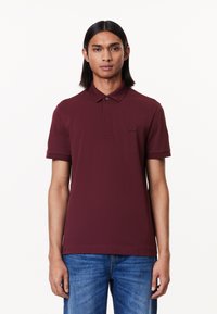 Burgundy polo skjorta med klassisk krage, korta ärmar och en subtil broderad logotyp. Tyget har en mjuk yta. Matchas med blå jeans.
