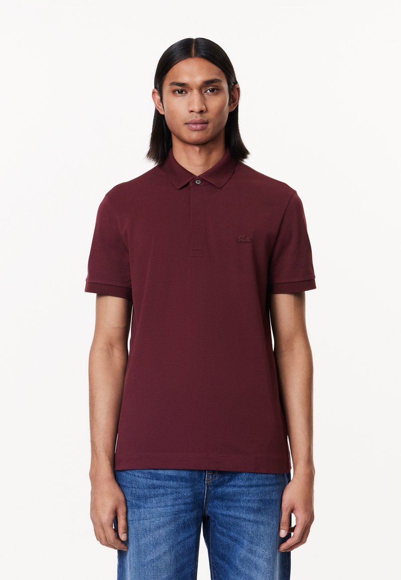 Burgundy polo skjorta med klassisk krage, korta ärmar och en subtil broderad logotyp. Tyget har en mjuk yta. Matchas med blå jeans.