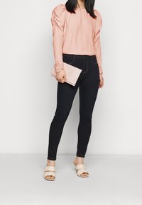 Růžová dlouhá rukávová halenka s nabíranými rameny, spárovaná s tmavými skinny džíny a taupe sandály. Drží světle růžovou kabelku.