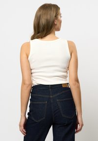 Vitt ribbat linne med rundad rygg, kombinerat med mörkblå högmidjade jeans som har två bakfickor och detaljer i guldstickning.