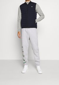 Námořnicky modrá prošívaná vesta přes šedé tričko s dlouhým rukávem, doplněná šedými tepláky se zelenými vyšívanými detaily a bílými vysokými teniskami.