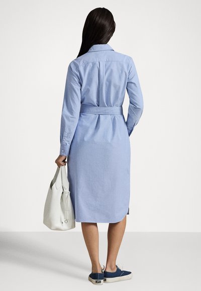 Kvinne med langt mørkt hår i lys blå kjole med belte som holder en hvit veske, iført marineblå sneakers, sett bakfra mot hvit bakgrunn.