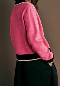 Femme portant un pull rose bordé de noir et blanc, debout de profil avec une main dans la poche d'une jupe ou d'un pantalon noir.