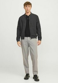 Chaqueta bomber negra con cierre frontal, llevada sobre un polo negro. Pantalones grises con pleats y zapatos negros con cordones. Texturas suaves en todas partes.