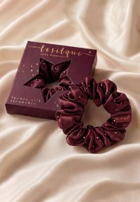 Donkerrode zijden scrunchie met kleine kraaltjes naast bijpassende merkdoos op beige satijnen stofachtergrond.