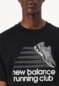 Juodas medvilninis marškinėlis su baltu grafiniu atvaizdu bėgimo bateliai ir tekstu "new balance running club" paryškintomis raidėmis.