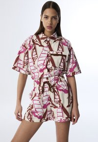 Camisa de manga corta con botones y pantalones cortos de cintura elástica en color crema, con un patrón en rosa y marrón de zapatos estilizados. Detalle de cuerda marrón.
