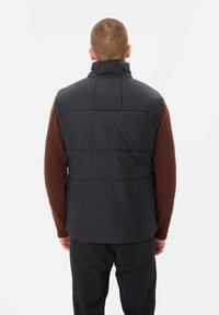Schwarze isolierte Weste mit hohem Kragen, horizontalen Nähten und glatter Oberfläche, über einem braunen Strickpullover getragen.