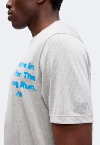 Homme en chemise perforée à manches courtes gris clair avec le texte bleu « We're In It For The Long Run » et le logo New Balance sur la manche.