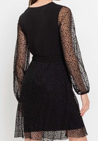 Svart klänning med en åtsittande liv och genomskinliga, långa ärmar med prickar. Texturerad tyg med något utställd kjol och slät midja.