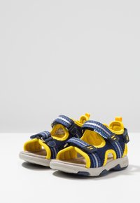 Nám blue a žluté dětské sandály s síťovým designem, nastavitelné popruhy a šedou gumovou podrážkou. Obsahují ochranu prstů a smyčku na patě.