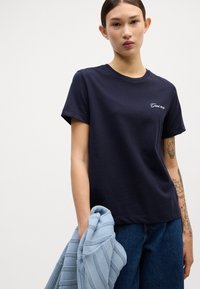 Mörkblå bomulls T-shirt med korta ärmar, som har broderad text. Modellen håller en ljusblå stickad tröja.