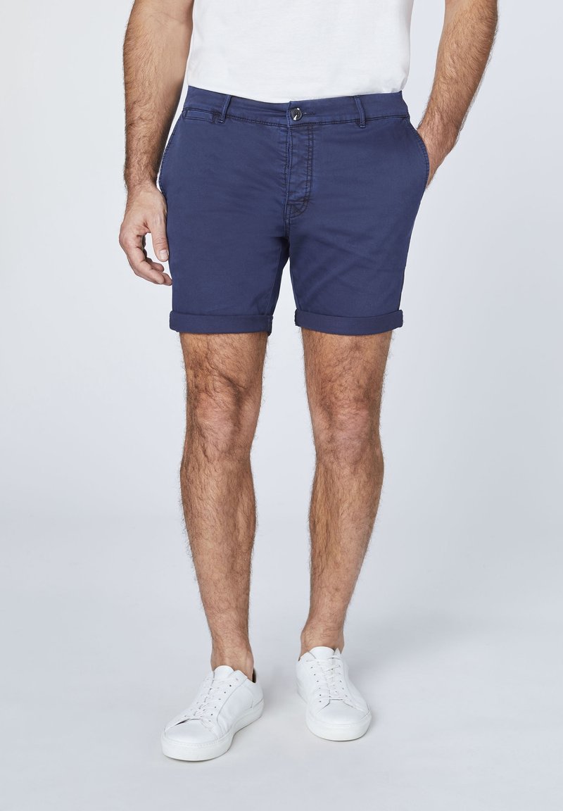 Homme portant un short bleu marine retroussé, des baskets blanches et un t-shirt blanc, debout avec une main dans la poche.