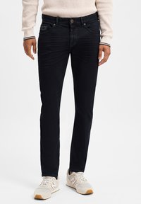 Un jean bleu foncé avec une coupe slim, une texture légèrement délavée et un design classique à cinq poches. Associé à des baskets beige clair.