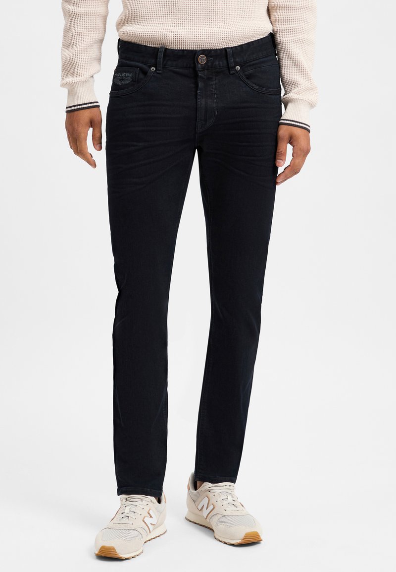 Un jean bleu foncé avec une coupe slim, une texture légèrement délavée et un design classique à cinq poches. Associé à des baskets beige clair.